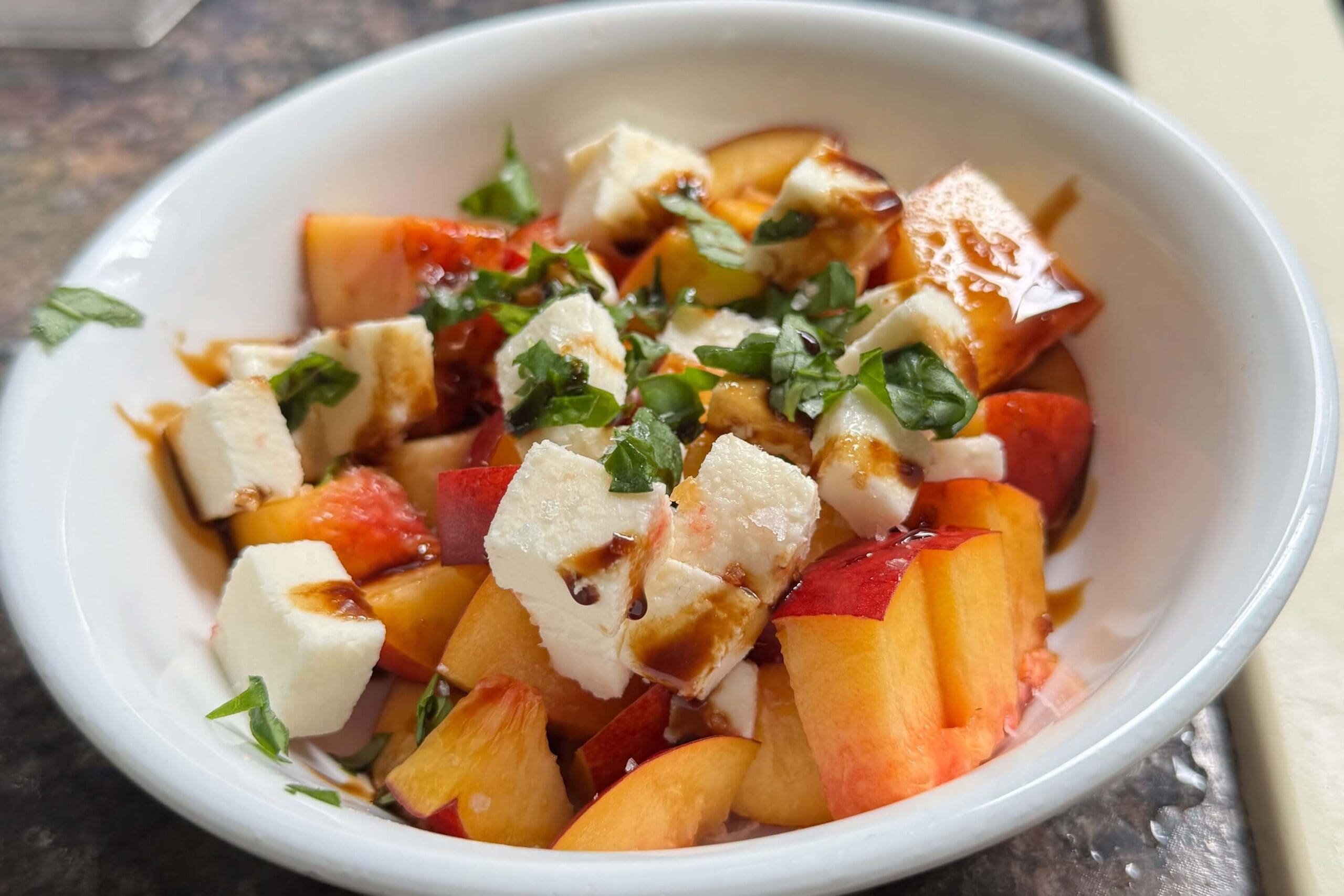 Close-up of nectarine salad with fresh mozzarella cubes, chopped basil, and balsamic glaze drizzled on top in a white bowl.