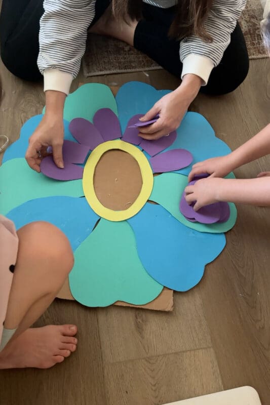 Arranging purple cardstock petals onto a blue and green cardboard base with an oval face opening to assemble a no sew flower costume for kids.