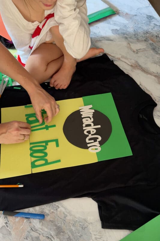 Hands arranging green foam letters that spell “Plant Food” while assembling a DIY costume t-shirt for a funny and creative Halloween outfit, with a child nearby helping in the process.