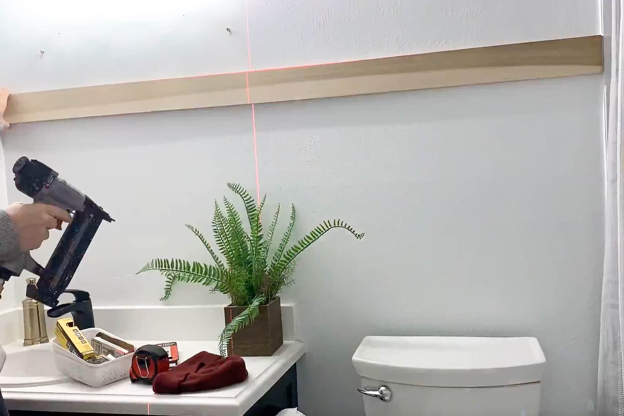 Installing the top rail for a DIY board and batten bathroom wall using a laser level and brad nailer above the vanity and toilet.