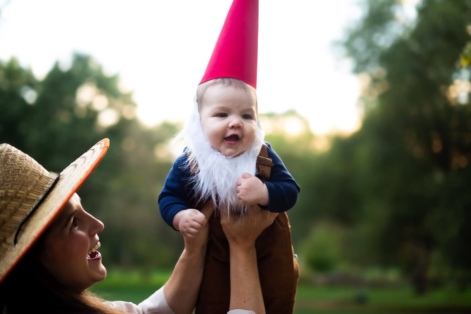 Baby in a DIY garden gnome costume—red cone hat and fluffy white beard—being held up by mom dressed as a gardener outdoors.
