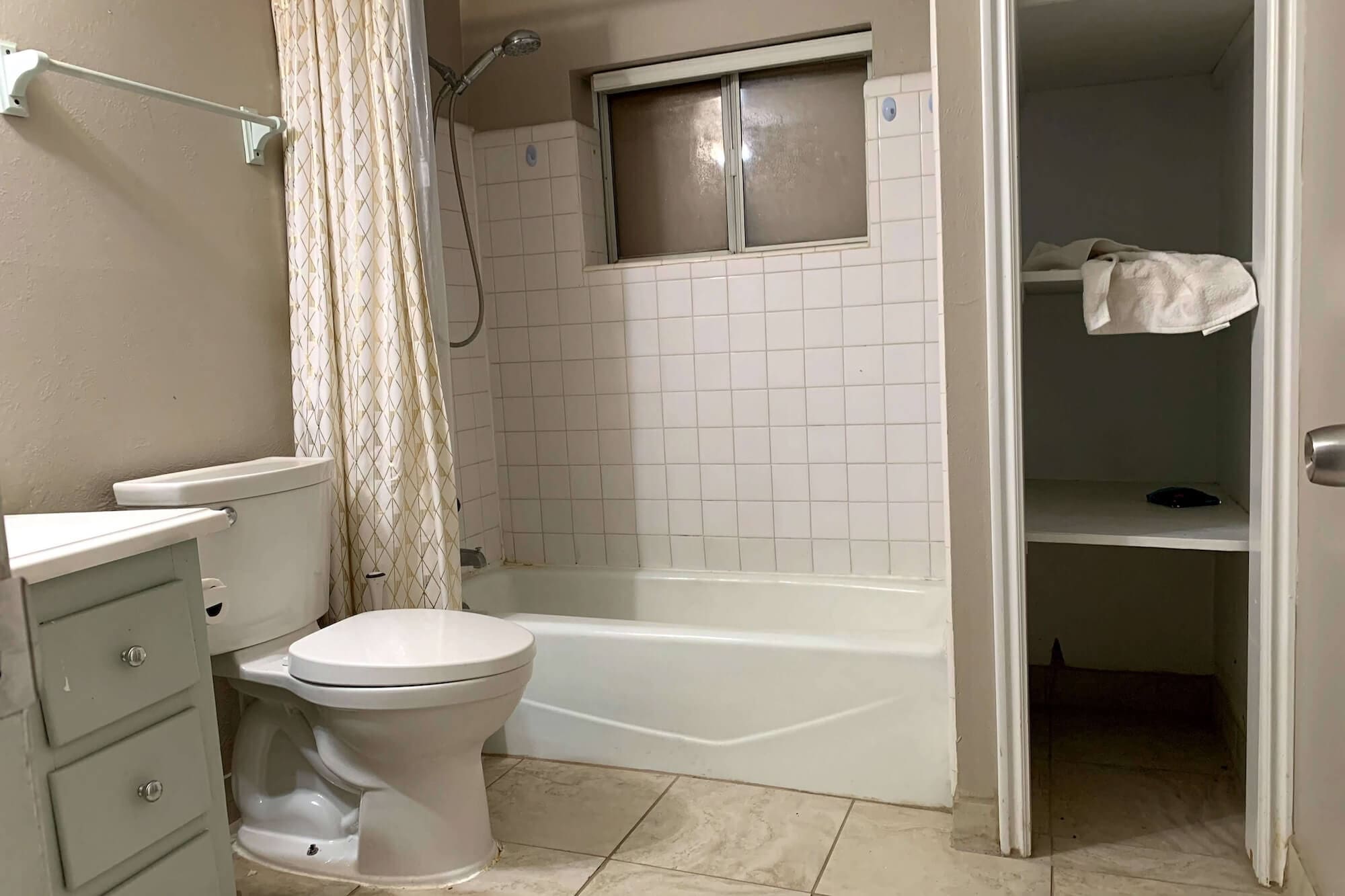 Before photo of small basement bathroom with beige tile shower surround, interior window, and wire shelves beside the tub.