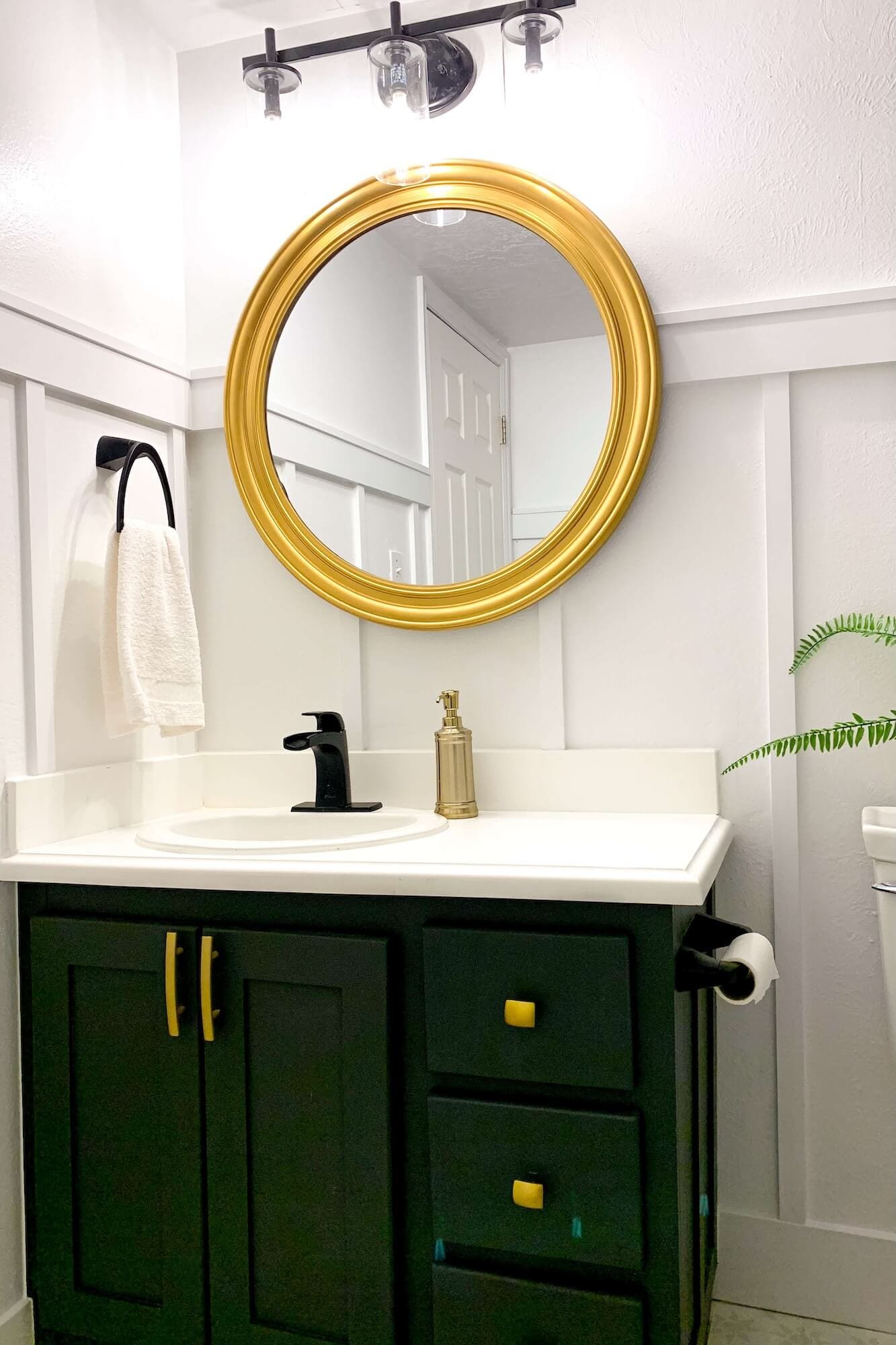 Small bathroom after a board and batten upgrade featuring a gold round mirror, black faucet, and dark green vanity.