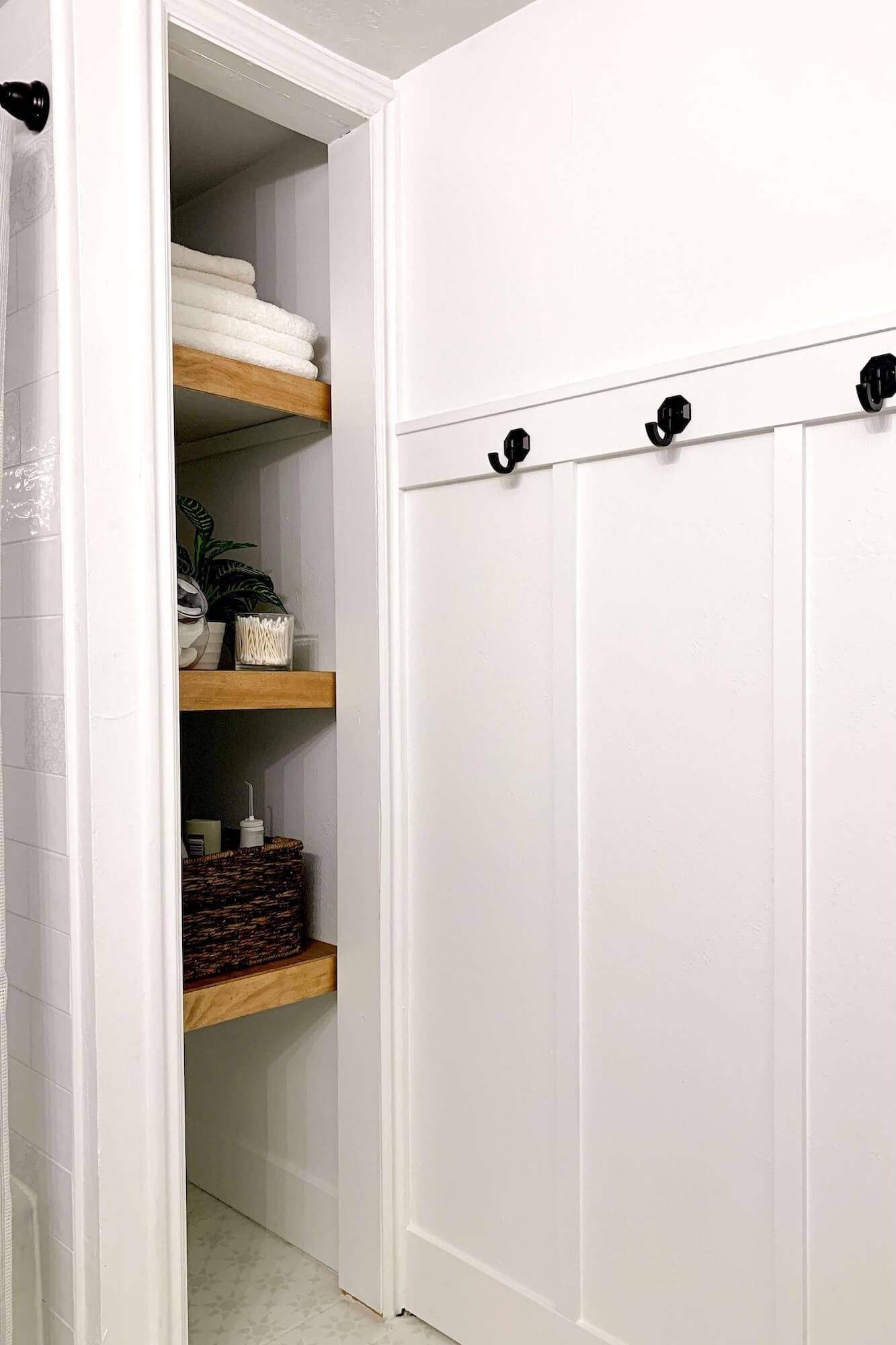 White board and batten bathroom wall with matte black hooks beside a shelving nook with wood shelves and rolled towels.
