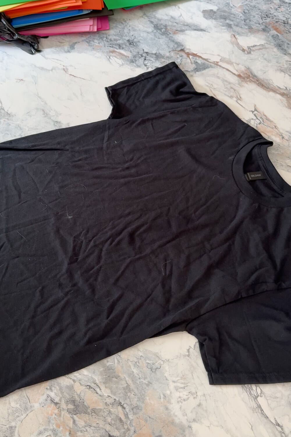 Plain black tee laid flat on a marble countertop with colorful craft paper sheets stacked in the background, ready for a DIY project.