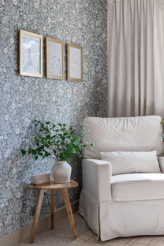 Blue floral wallpaper wall with a cream armchair, linen curtains, three gold frames, and a plant on a wood stool.