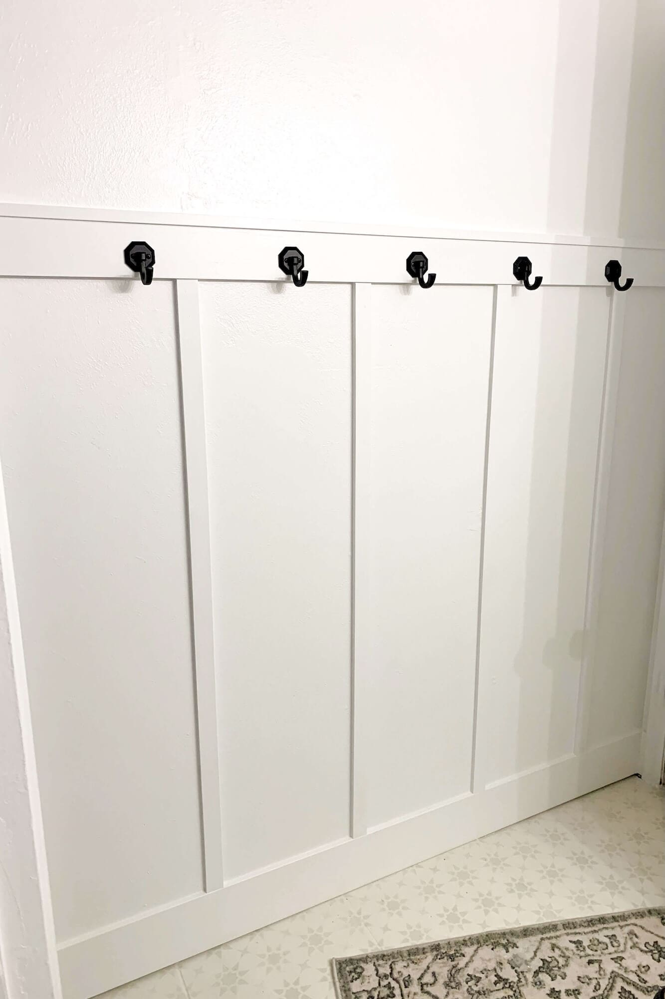 DIY board and batten bathroom accent wall painted white with evenly spaced black coat hooks along the top rail.