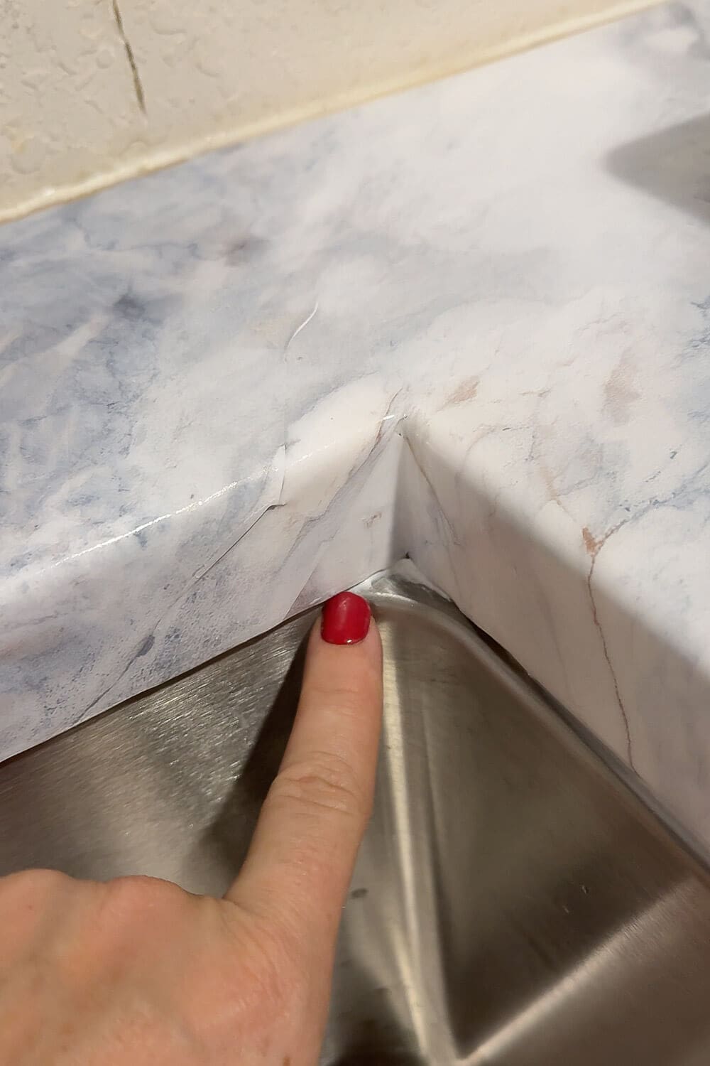 Inside sink corner of a kitchen counter wrapped in marble contact paper, finger pointing to a small seam/wrinkle—example of trimming and smoothing for faux marble countertops.