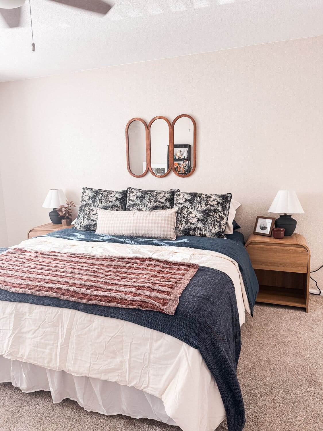 cozy fall bedding in a guest room refresh featuring navy quilt, cream duvet, rust knit throw, plaid lumbar, wood nightstands with lamps, and a triple wood mirror above the bed