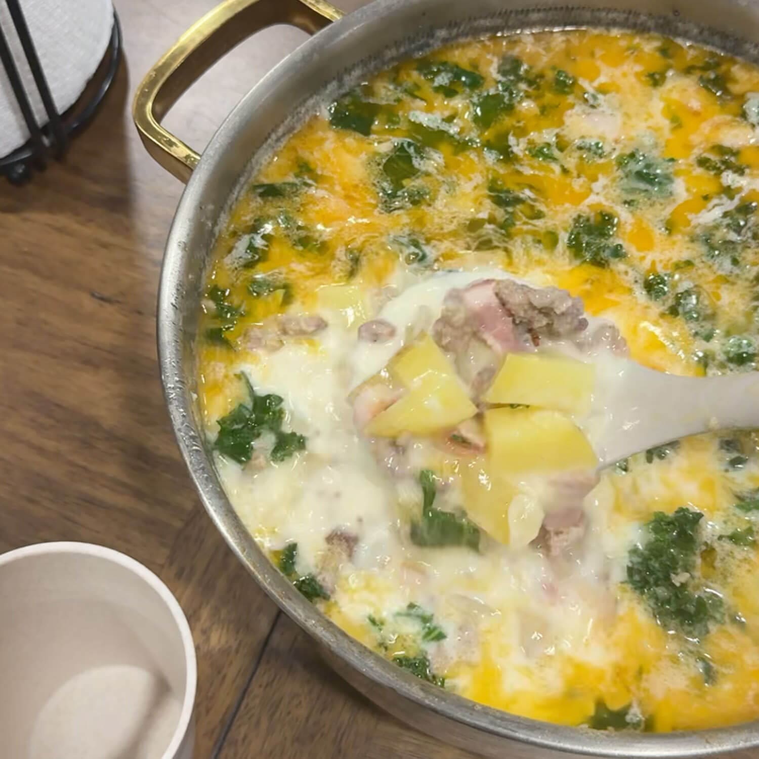 Creamy homemade Zuppa Toscana in a gold-handled pot, with a ladle lifting Yukon gold potatoes, Italian sausage, bacon, and kale in a rich garlic-cream broth.