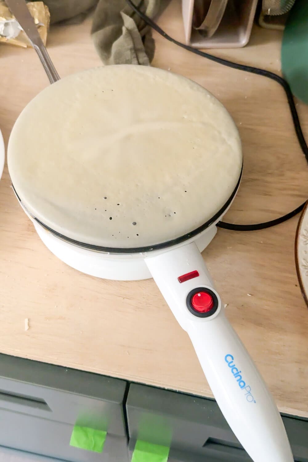 Cordless crepe maker cooking a thin batter layer on the nonstick plate, red power light on—step for making fast, thin café-style crepes at home.