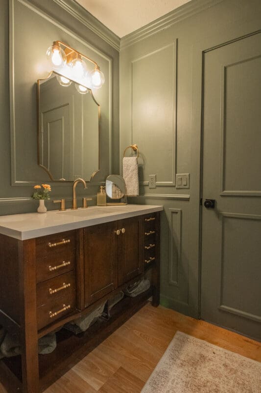 Dark green bathroom with picture frame molding, brass mirror and light, and a wood vanity with a white countertop.