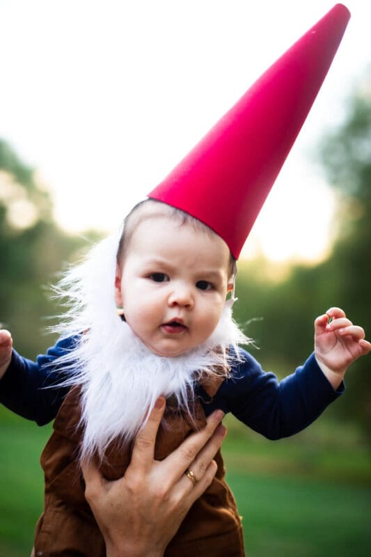 Baby in a dressed as a garden gnome for Halloween is wearing a tall red cone hat, fluffy white beard, navy shirt, and brown overalls outdoors.