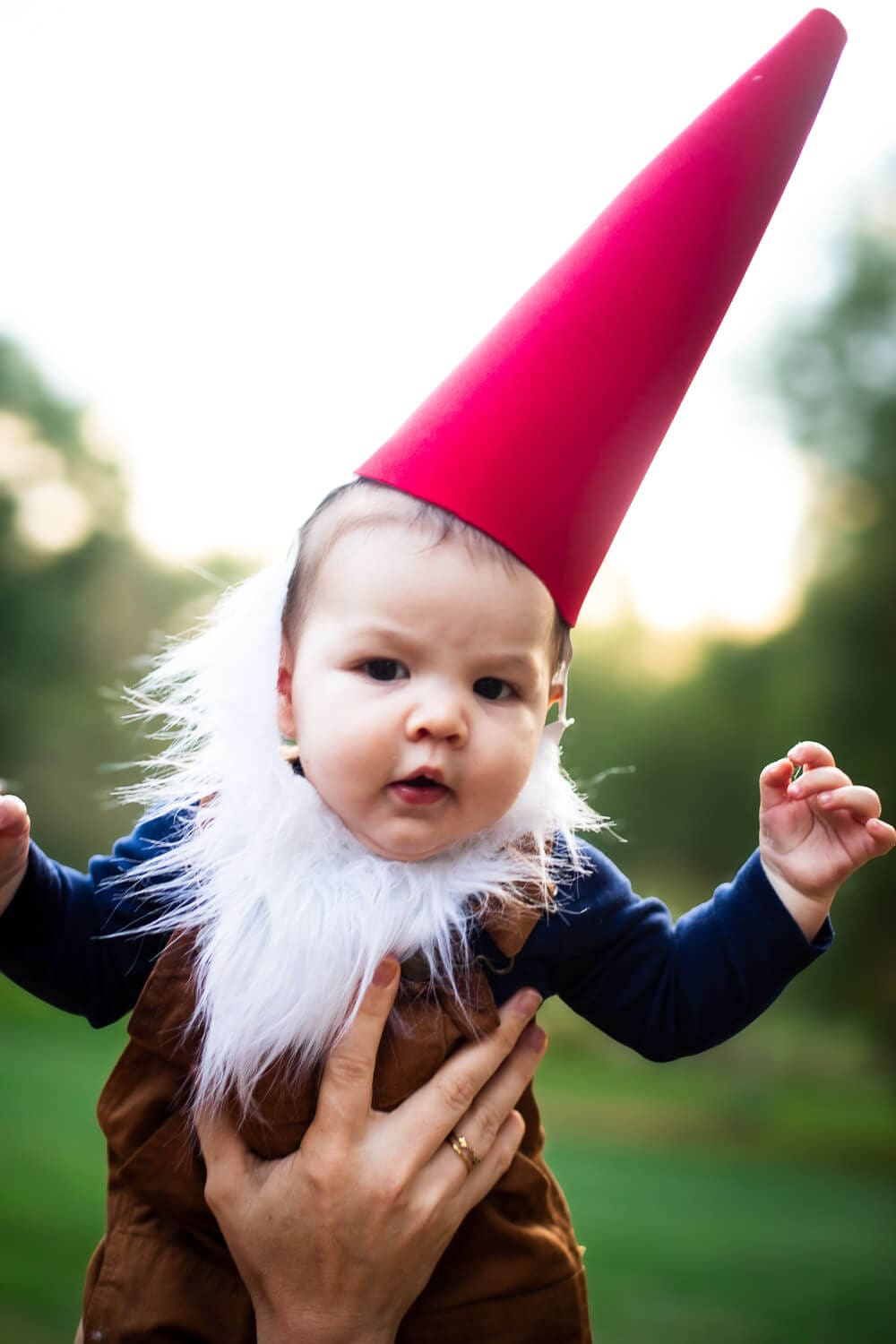 Baby wearing a red cone hat, faux fur beard, and brown overalls as part of a DIY garden gnome costume for Halloween.
