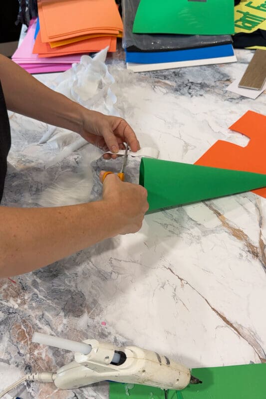 Securing elastic between the white faux fur beard and green foam hat using hot glue for an easy no-sew Halloween look.