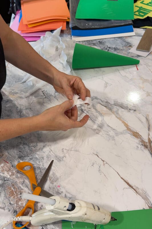 Gluing a white elastic strap inside a green foam gnome hat to attach it to the faux fur beard for a DIY kids last-minute Halloween look.