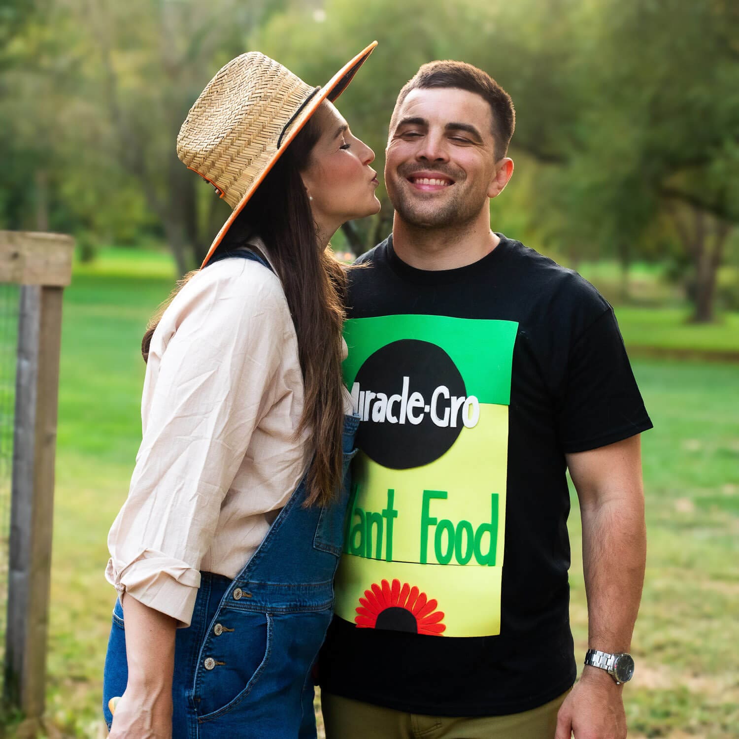 Couple dressed in matching DIY Halloween costumes standing outdoors — wife in gardener overalls and hat, husband wearing a funny costume t-shirt designed to look like a plant food bag.
