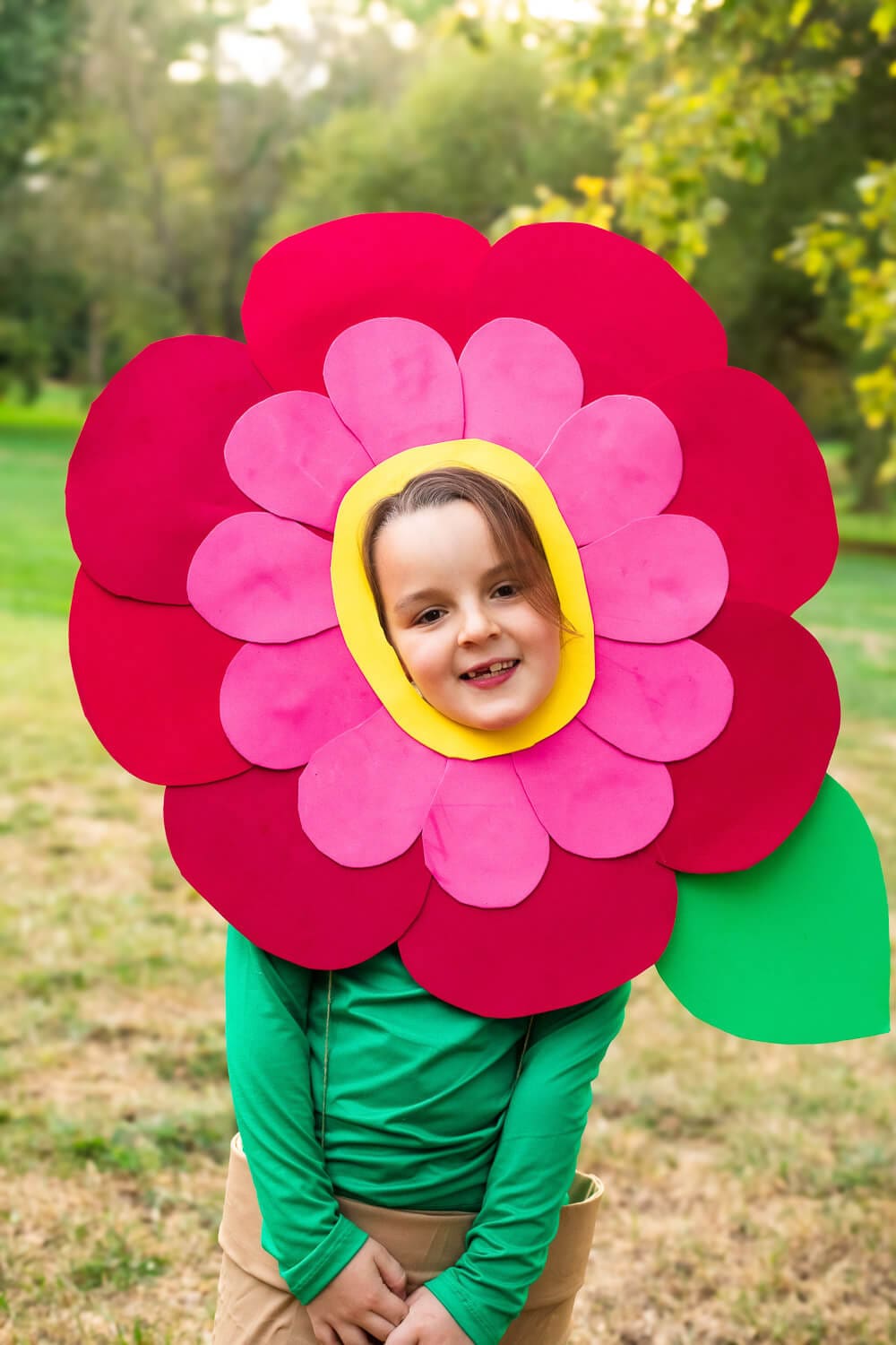 Child in a DIY flower costume with layered red and pink petals and a yellow center, wearing a green “stem” shirt and cardboard flower pot skirt, smiling outdoors.