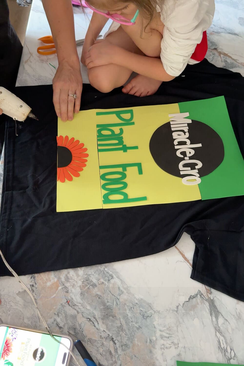 Hands gluing the final foam pieces onto a black shirt with a child watching, finishing a colorful DIY costume t-shirt for a funny homemade Halloween idea.