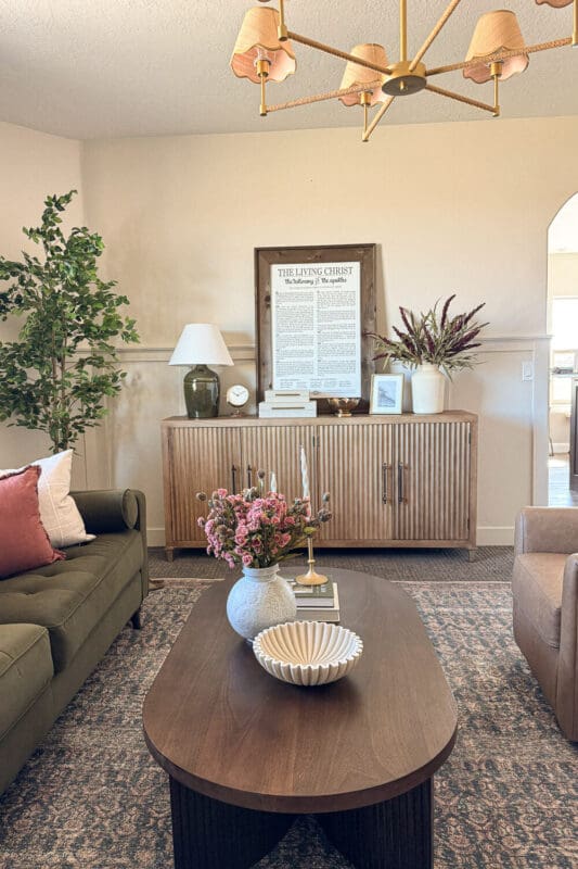 Formal living room after with brass chandelier, wood slatted credenza, table lamp, framed art, faux tree, and layered rug