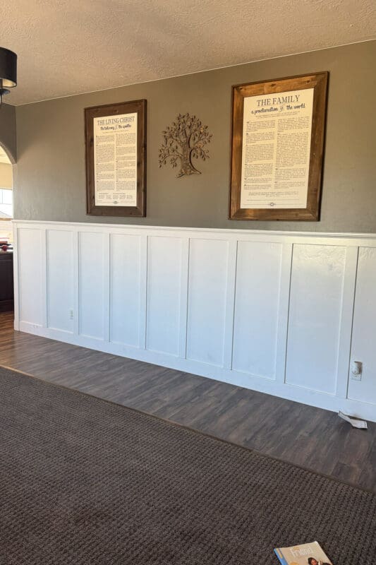 Front sitting room before with tall white board and batten on gray wall, wood-look floors meeting gray carpet, framed artwork, and open wall awaiting seating.