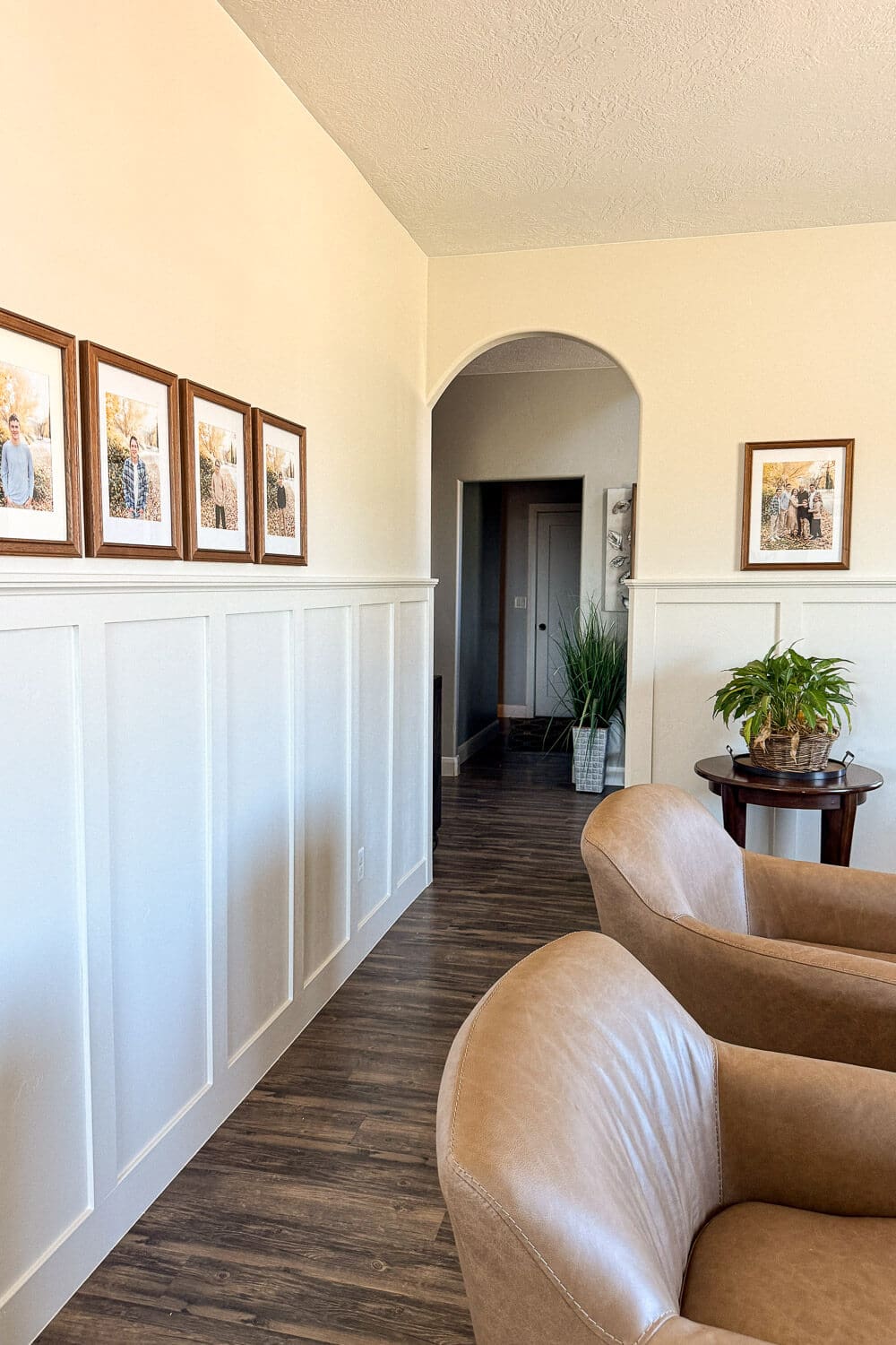 Formal living room wall with white board and batten, arched doorway, family photo gallery, wood floors, and tan leather chairs in the foreground.