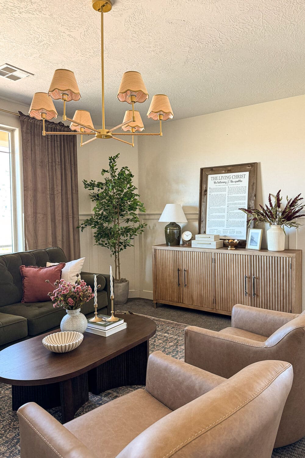 Formal living room with brass chandelier, tan leather chairs, green velvet sofa, faux tree, fluted wood credenza, and styled oval coffee table.