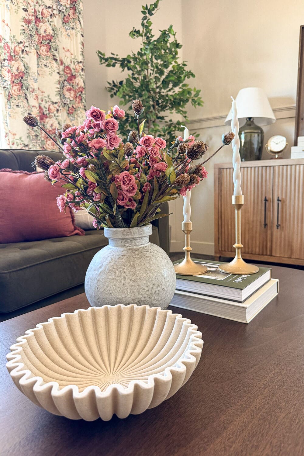 Coffee table styling in a formal living room with fluted bowl, vase of pink flowers, brass candlesticks on books, green sofa, floral curtains, and faux tree.