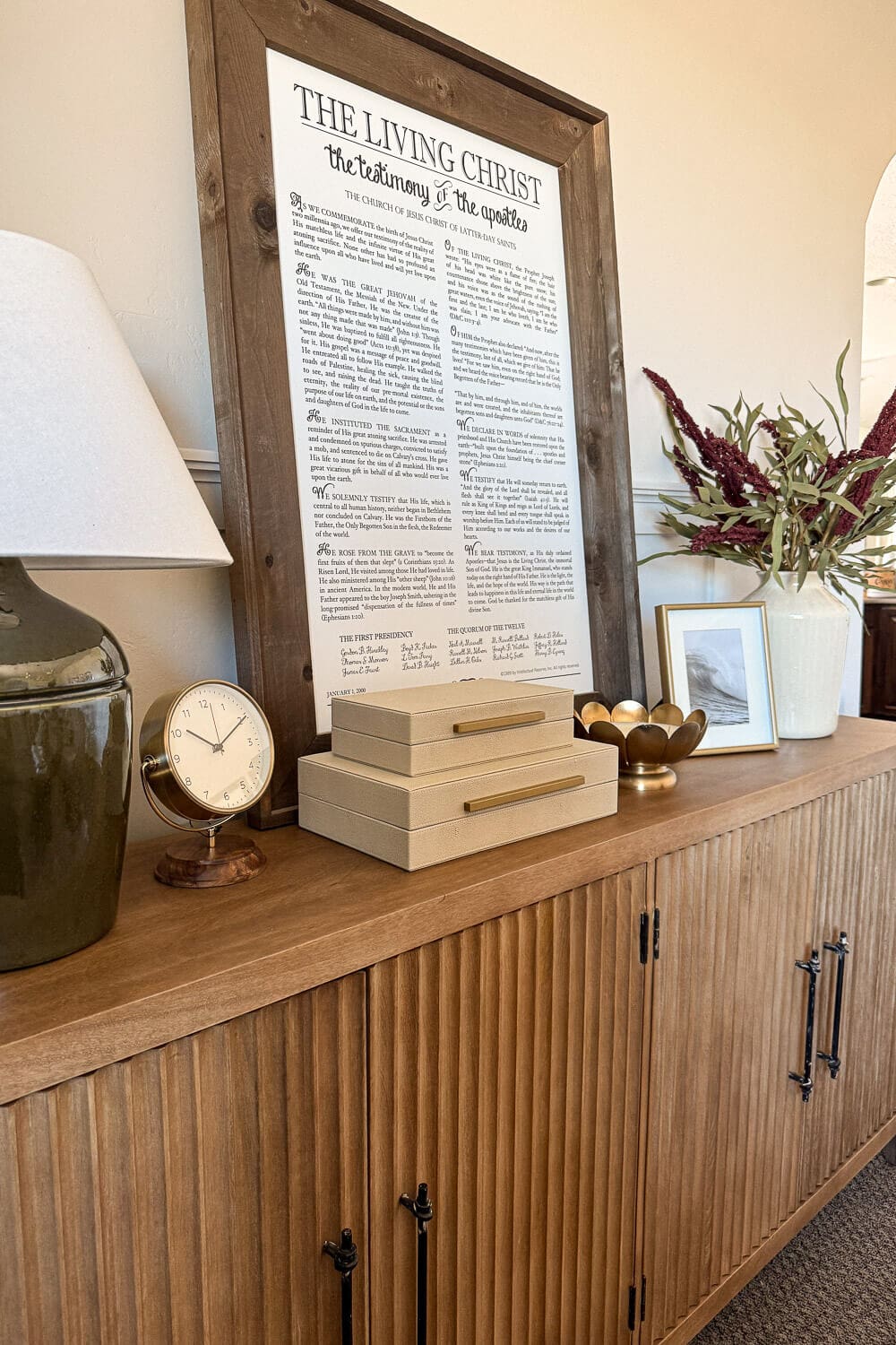 Console table decor in a formal living room with green ceramic lamp, small clock, stacked storage boxes, framed art, and vase with stems on a slatted wood credenza.