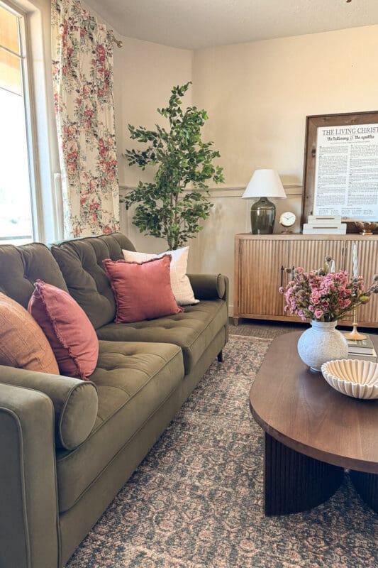 Formal living room with floral curtains, green velvet sofa, pink pillows, faux tree, ribbed wood credenza, and styled coffee table showing how brighter drapery lightens the space.