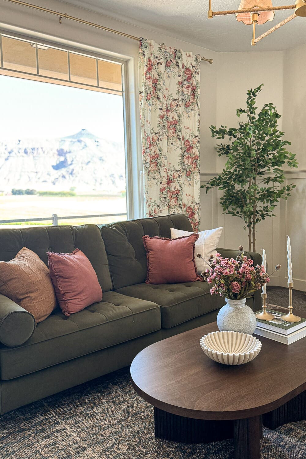 Green velvet sofa with pink pillows beside floral curtains and a large window with mountain views, styled coffee table in the foreground.