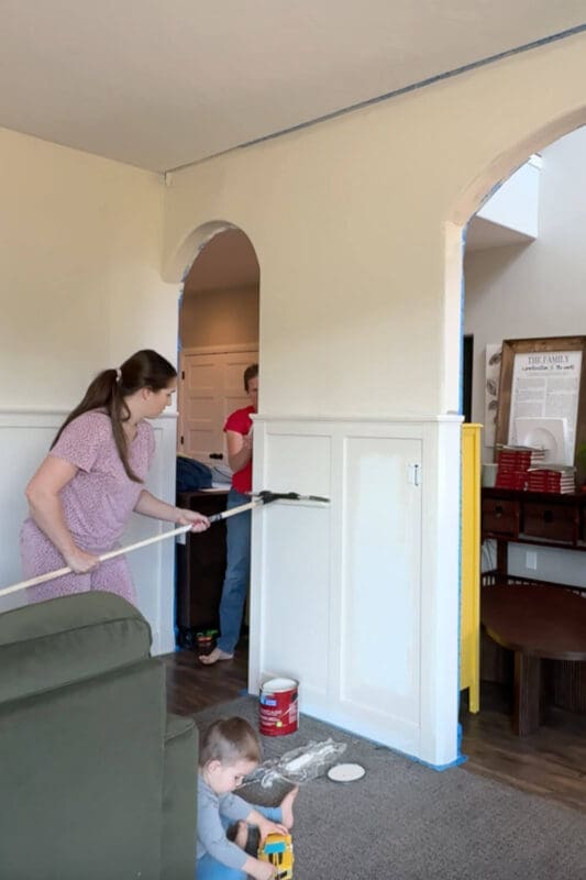 Painting a front room in Sherwin-Williams Accessible Beige with an 18-inch roller; board and batten walls prepped with tape while a child plays nearby.
