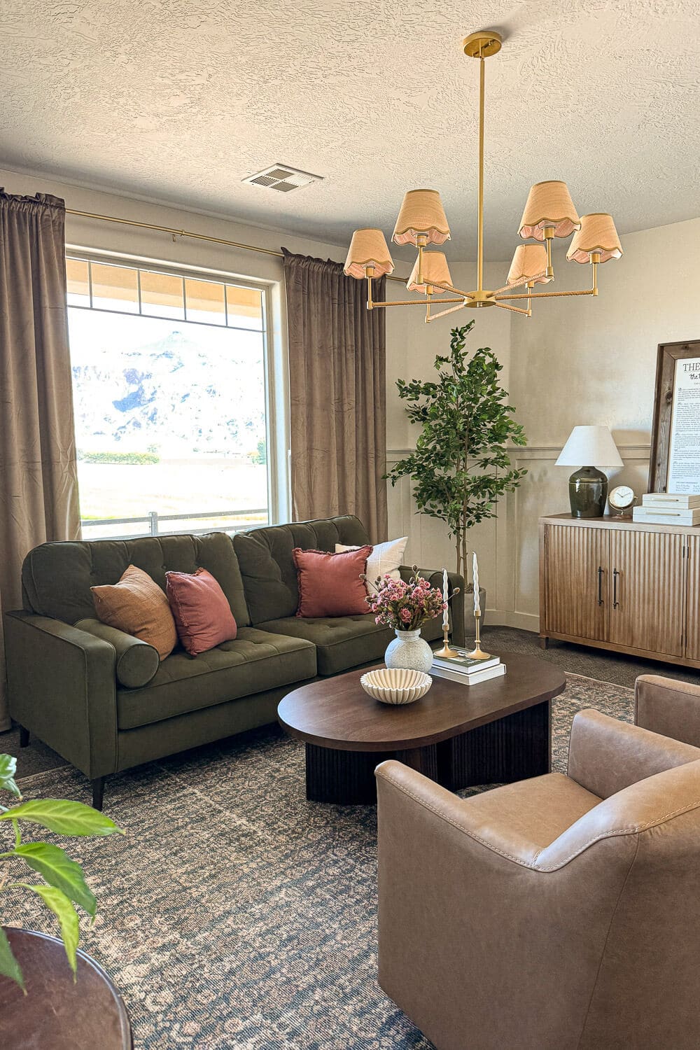 Cozy formal living room seating area with green tufted sofa, blush pillows, leather club chairs, vintage-style rug, wood credenza, and tall faux tree for a conversation layout.