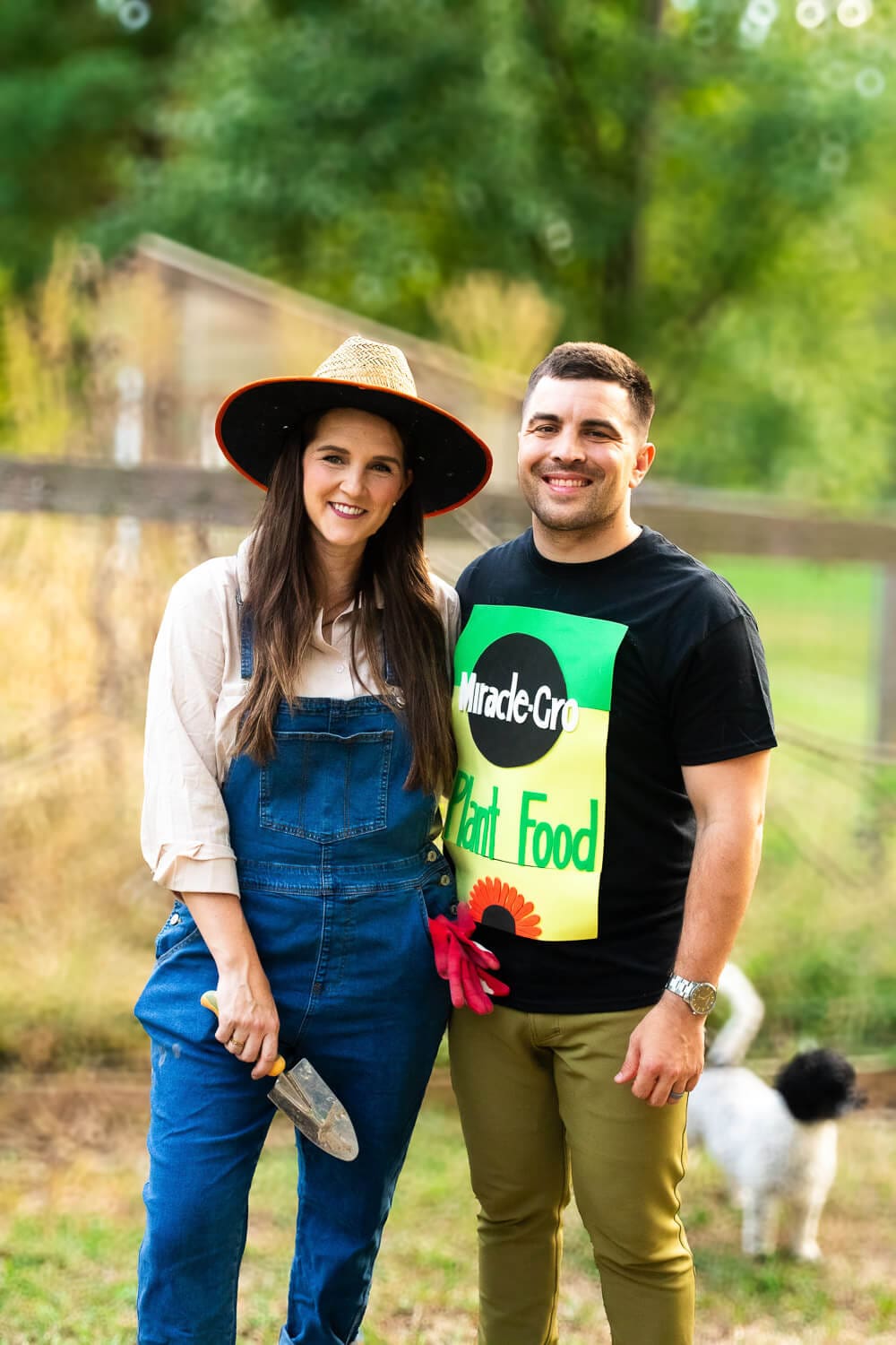 Couple in garden-themed DIY Halloween costumes: woman dressed as a gardener in overalls and straw hat holding a trowel, next to man wearing a Miracle-Gro plant food T-shirt.