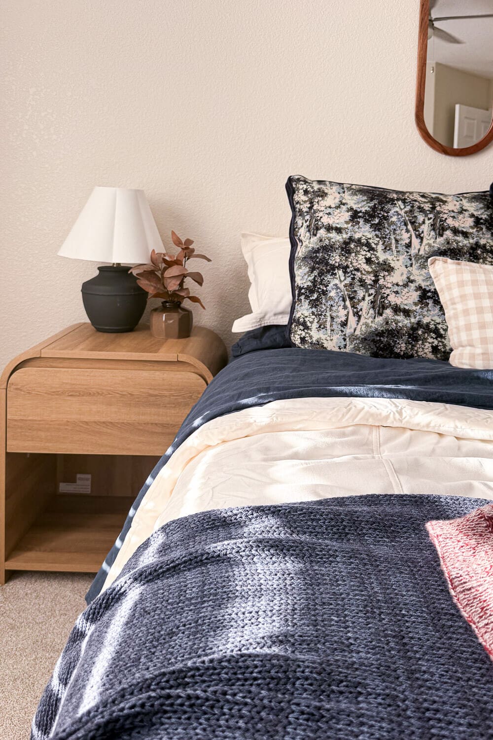 wood nightstand styled with black lamp and small vase beside patterned euro pillow; navy quilt, cream duvet, and knit throw layered for a cozy fall bedding guest room refresh