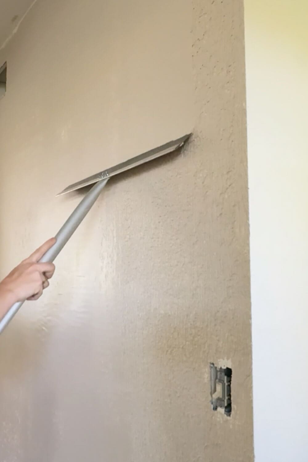 How to skim coat a wall: close-up of a magic trowel smoothing wet joint compound over textured drywall near an outlet.