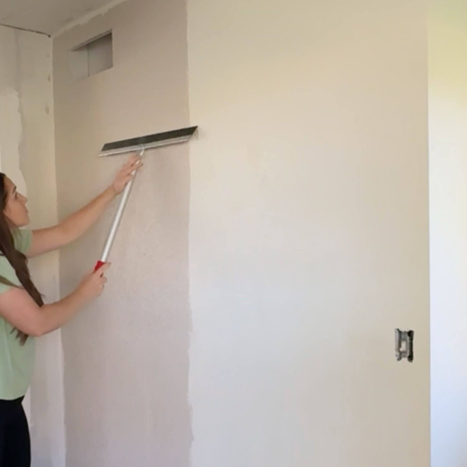 How to skim coat a wall to remove texture; woman using a magic trowel to pull joint compound smooth on a bedroom wall.