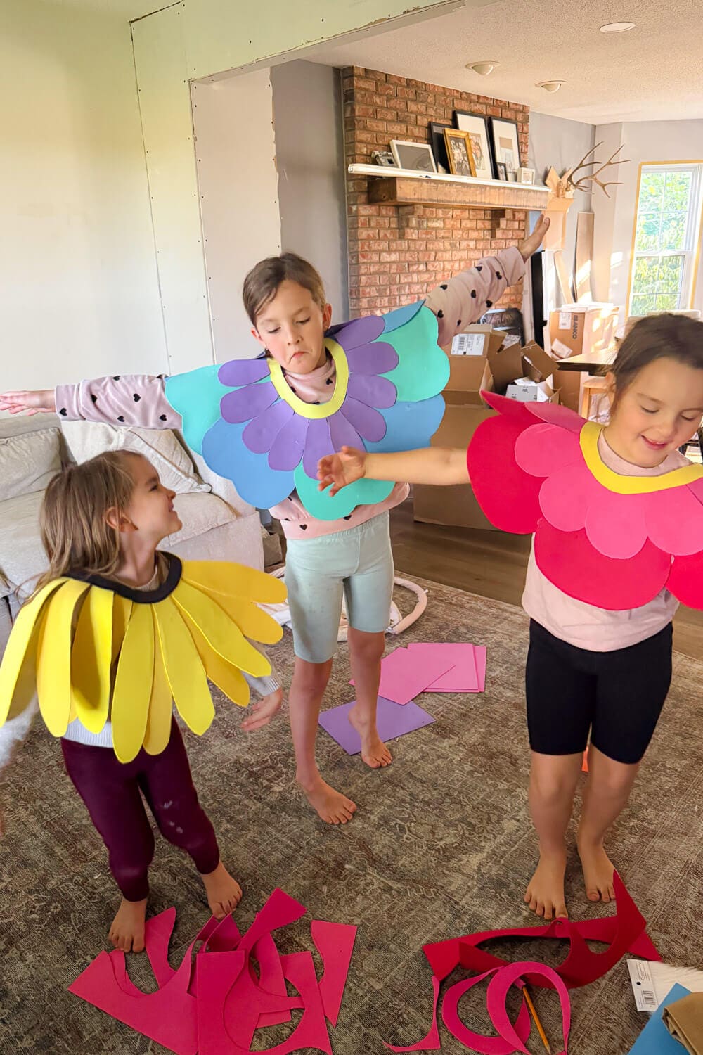 Kids trying on no sew cardstock flower costume collars—yellow sunflower, blue and purple daisy, and red and pink petals—with paper scraps on the floor during a DIY test fit.