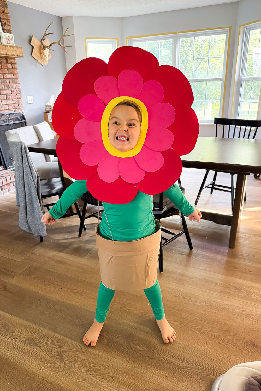Child modeling a no sew cardstock flower costume with layered red and pink petals, yellow face ring, green stem outfit, and a brown flower-pot skirt—easy DIY Halloween costume for kids.