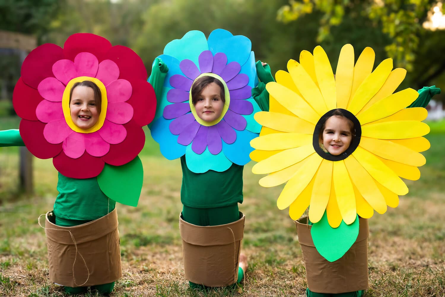 Three kids in DIY no-sew flower costumes: red, blue-purple, and yellow sunflower petal headpieces with green “stem” outfits and cardboard flower-pot skirts.