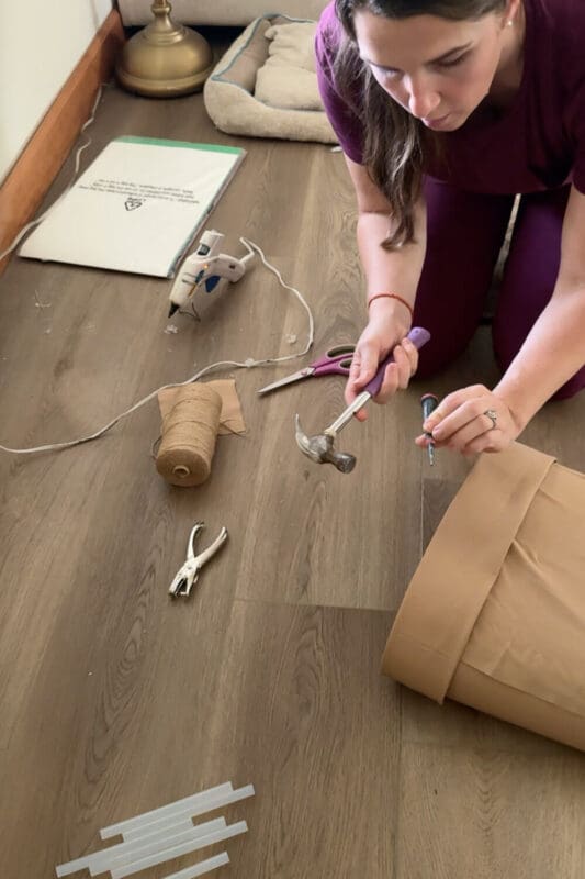 Punching holes in the rim of a brown fabric plant-pot skirt using a screwdriver and hammer to add straps for wearing.