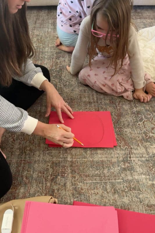 Mom tracing a large petal shape onto stacked red cardstock while her daughter watches, first step in a no sew cardstock flower costume for kids.