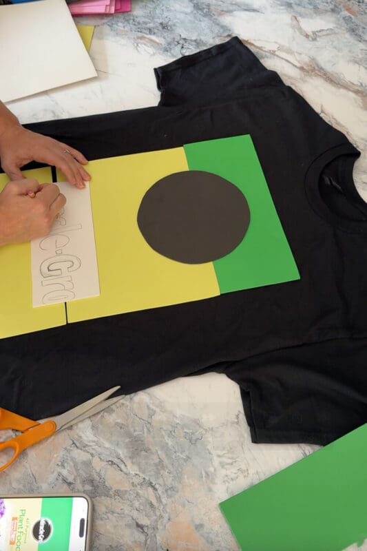 Tracing letters onto white craft foam beside a black, yellow, and green layout while creating a DIY costume t-shirt design for an easy, funny Halloween project.
