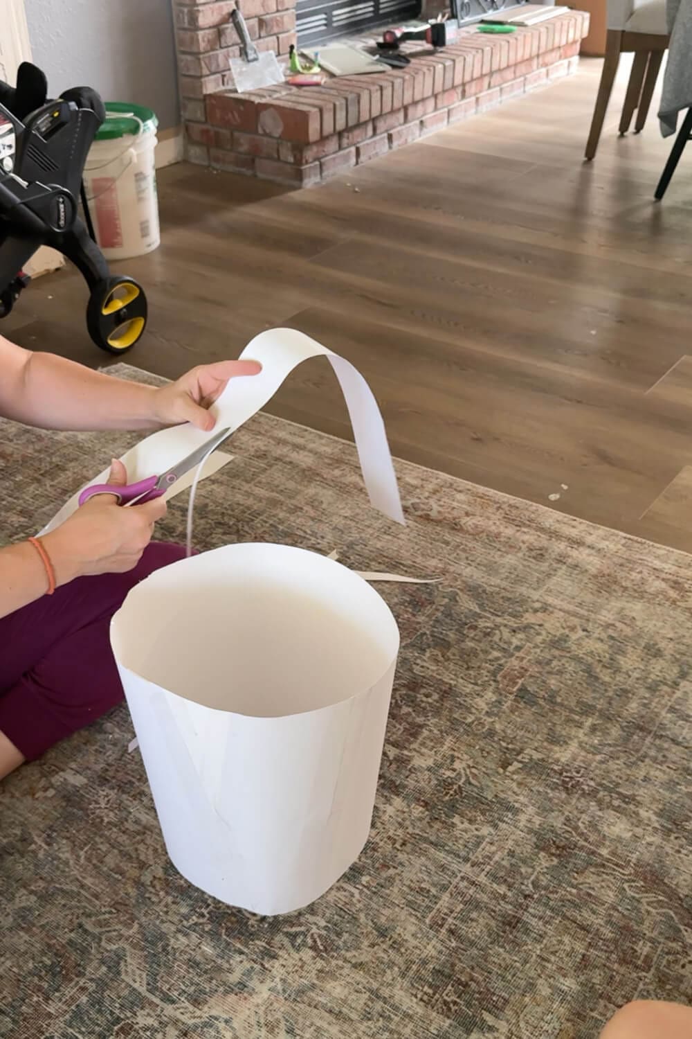 Trimming a long strip of white poster board with scissors next to a formed cylinder—shaping the flower-pot skirt for a no-sew cardstock flower costume.