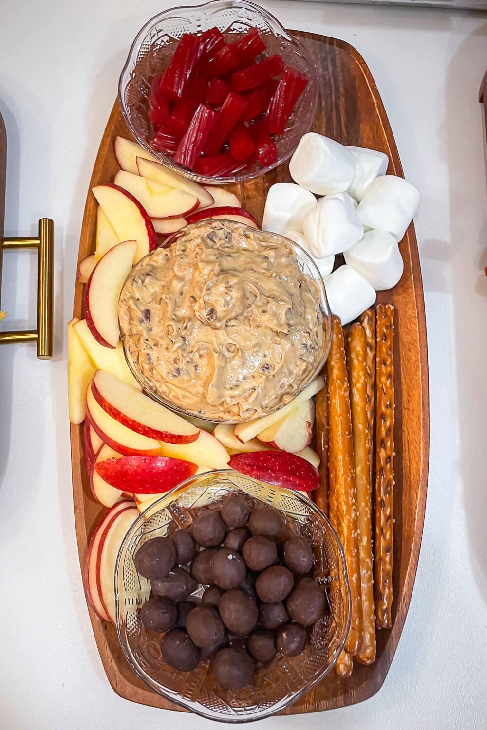 Overhead view of a long wooden dessert board with cream cheese apple dip in the center, sliced apples, marshmallows, pretzel rods, red licorice pieces, and a bowl of chocolate candies.