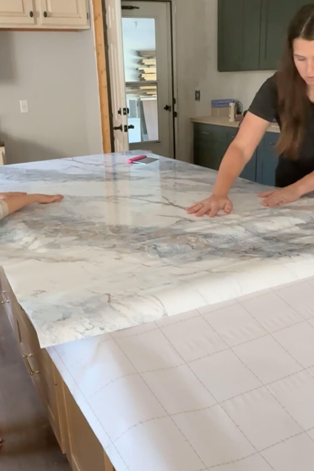 Smoothing a large sheet of marble contact paper over a plywood kitchen island to create DIY countertops that look like marble before sealing with epoxy.