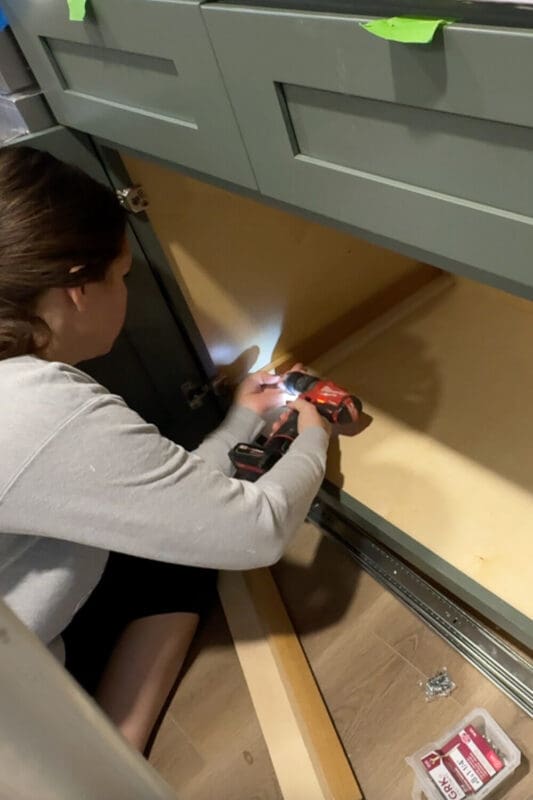 Woman crouched inside a lower kitchen cabinet, screwing drawer slide hardware to the cabinet walls to prep for a slide-out pots and pans storage organizer.