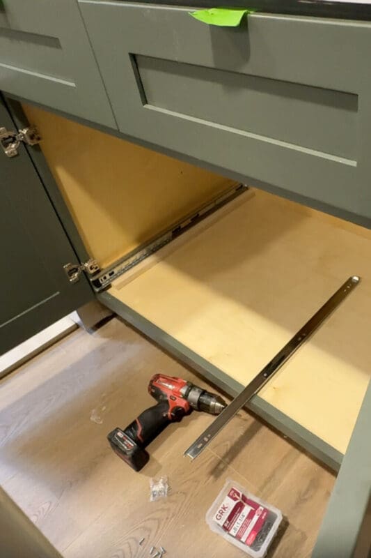 Empty lower cabinet with freshly installed full-extension drawer slides and a drill on the floor, ready for the custom pots and pans storage drawer box.