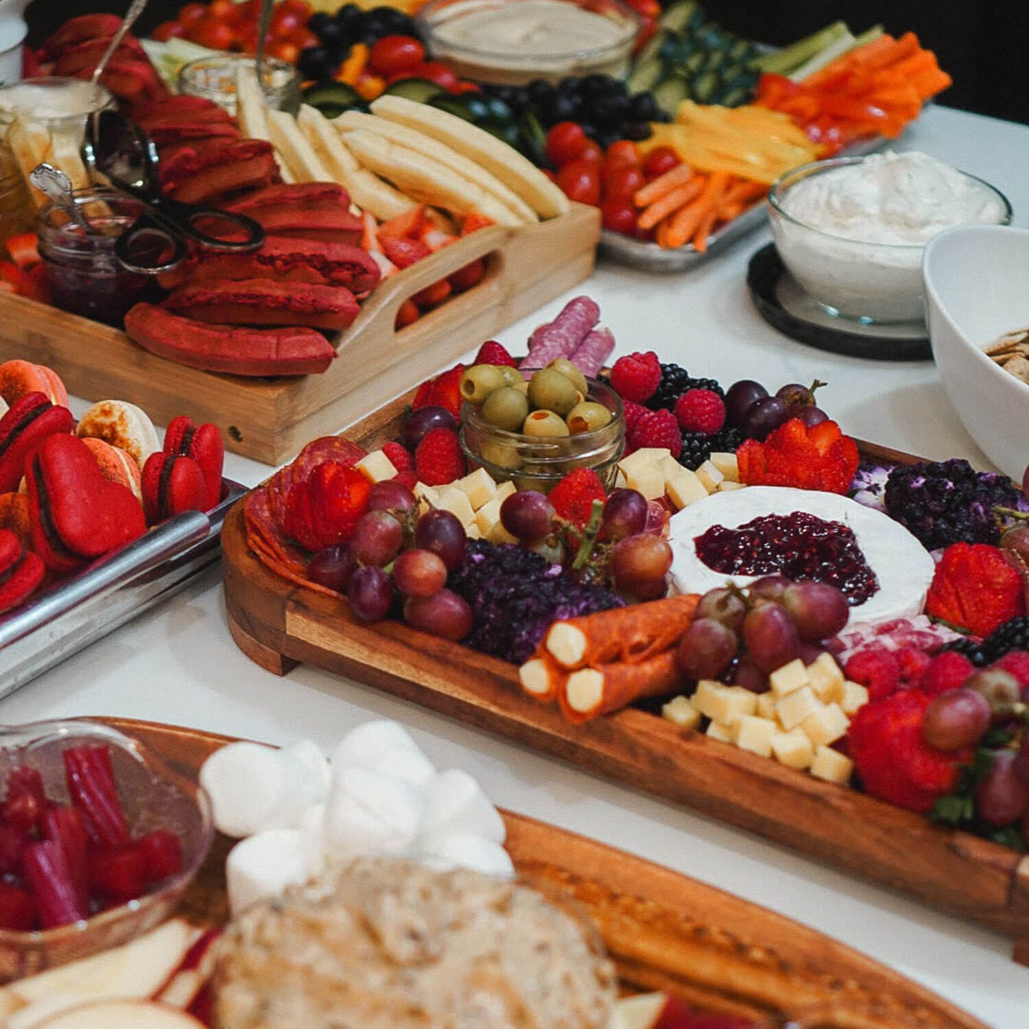 Charcuterie board ideas for a party—a table of boards with brie topped with jam, grapes, olives, salami, cheese cubes, red macarons, waffles, and a veggie tray with dips.