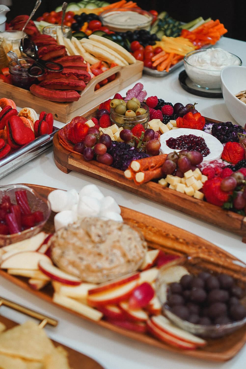 Charcuterie board ideas for a party: a buffet of boards with brie topped with jam, grapes, olives, cheese cubes, red macarons, apple slices with sweet dip, and a veggie tray in the background.